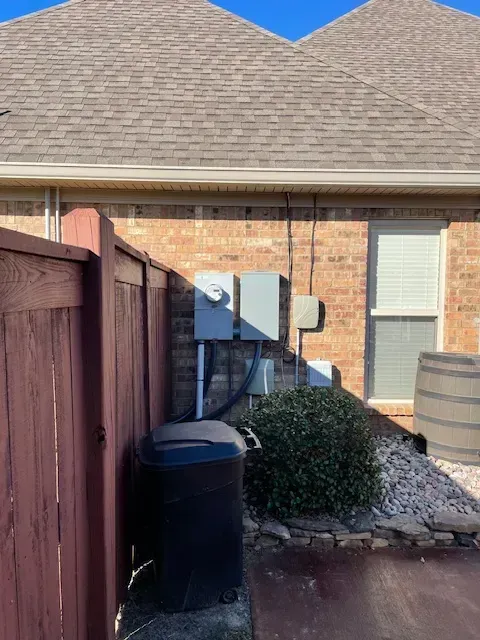 Electrical equipment attached to a brick wall next to a fence and a trash can, under a roof.